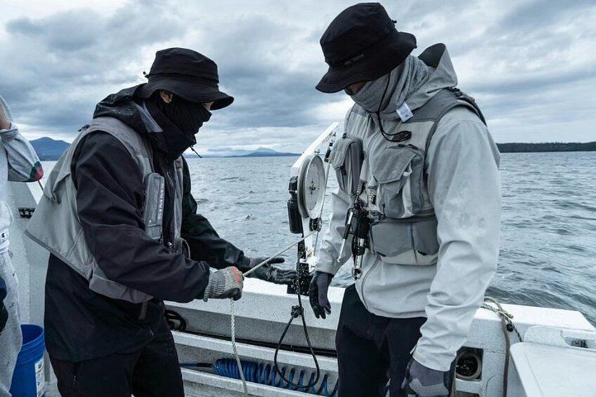 Dos personas en un bote, una con sombrero y chaqueta negra, la otra con chaqueta blanca, con un cuerpo de agua y montañas al fondo. 3