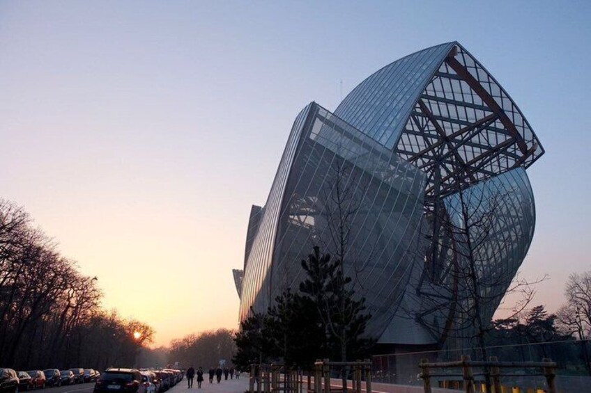A modern glass building with a unique curved structure, surrounded by trees and parked cars. 2