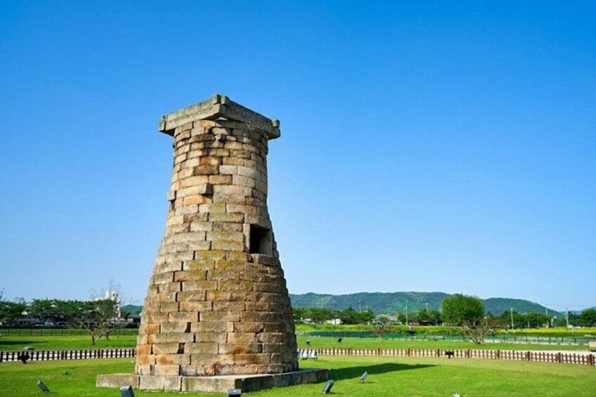 A stone tower with a flat top, surrounded by a grassy field and a fence, with hills in the background. 3