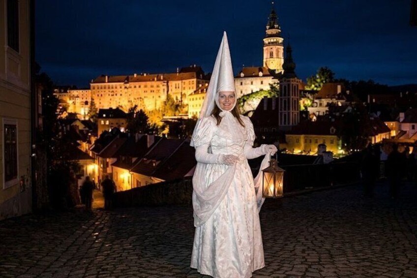 A person in historical attire standing on a cobblestone street with illuminated buildings and a tower in the background. 3