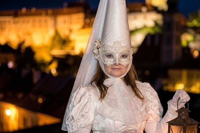 A person in a white costume with a cone hat and a lantern, standing in front of a lit-up building at night. 1