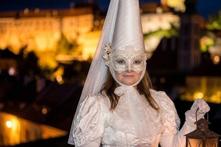 Eine Person in einem weißen Kostüm mit spitzen Kopfbedeckung hält eine Laterne und steht vor einem beleuchteten Gebäude bei Nacht. 1