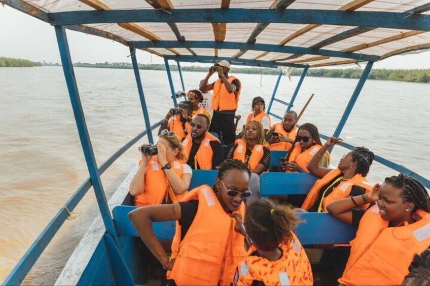 Een groep mensen in een boot, gekleed in oranje reddingsvesten, met een rivier en groen in de achtergrond. 1