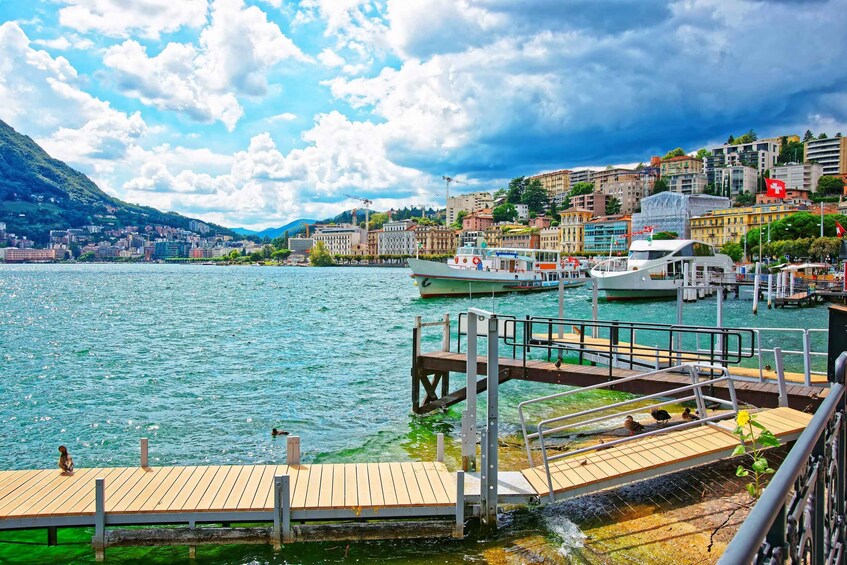A waterfront with a wooden dock, boats, and a cityscape with buildings and a mountain in the background. 1