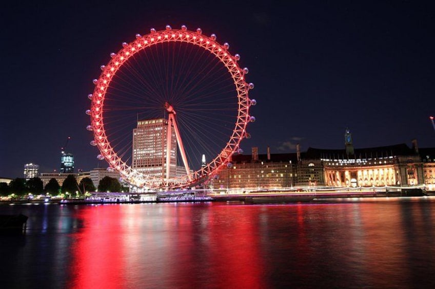 A large Ferris wheel at night with reflections on the water, a lit-up building, and a bridge in the background. 2