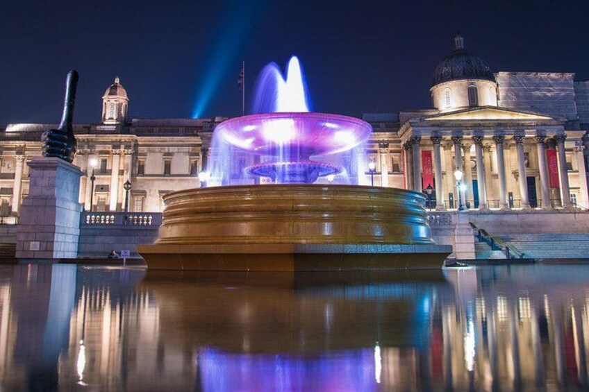 Ein Brunnen mit in Blau und Violett beleuchteten Wasserstrahlen, deren Spiegelung auf einer ruhigen Wasseroberfläche erkennbar ist. Im Hintergrund ein klassizistisches Gebäude mit Säulen und einer Kuppel. 3