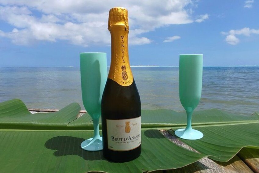Una botella de champán Brut Jaoua sobre una mesa con dos copas verdes, frente a un telón de fondo de playa y océano. 2