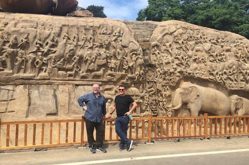 Two men standing in front of a large rock with detailed carvings, including figures and animals. 4