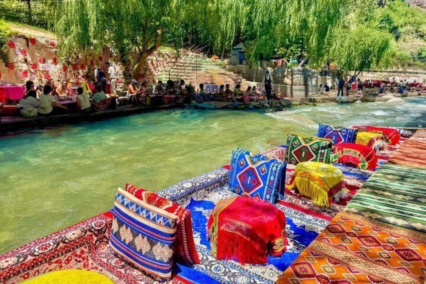 Un río con barcos decorados con telas coloridas, gente sentada y de pie a lo largo de la orilla, y árboles al fondo. 1