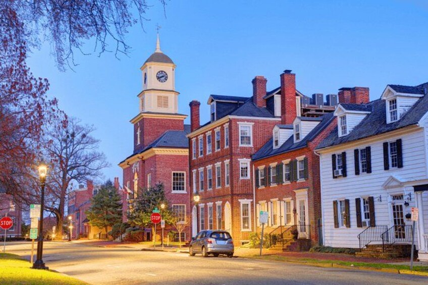 A historic town square with a prominent clock tower, red brick buildings, and a white building with black shutters. 1