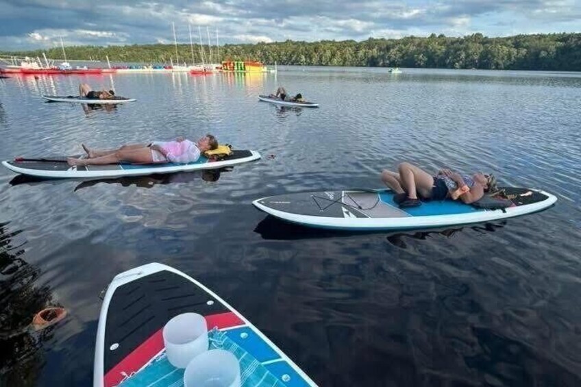 People lying on surfboards in calm water, surrounded by boats and a forested shoreline. 4