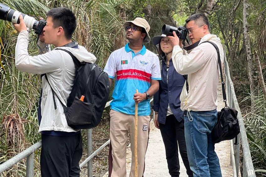 Eine Gruppe von Menschen auf einer Brücke, einige mit Kameras, eine Person mit Wanderstock, und eine weitere Person trägt ein T-Shirt mit der Aufschrift 'BAIGLADESH'. 1