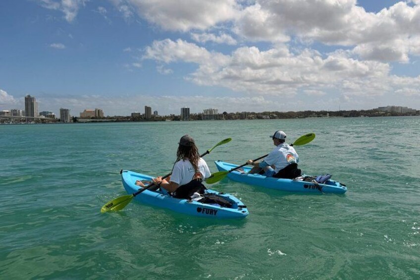 Two people kayaking on a calm body of water with a city skyline in the background. 5