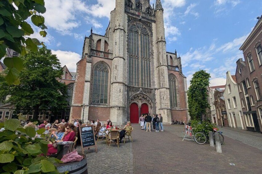 A historic church with a tall tower and large windows, surrounded by a cobblestone square with people sitting at outdoor tables. 1