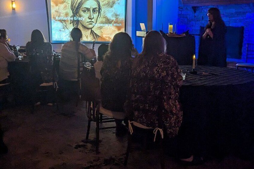 A dimly lit room with people seated at tables, a projection screen displaying a portrait, and a person standing at the front. 3