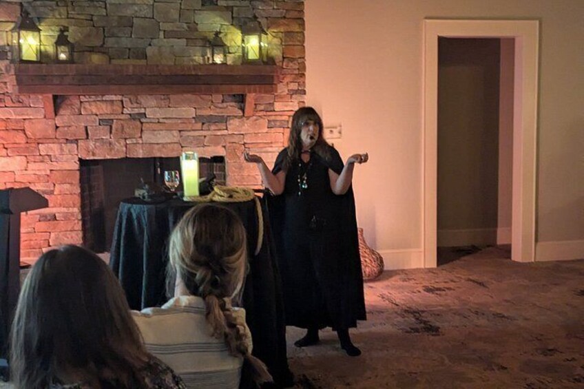 A person in a black dress standing in a room with a stone fireplace, a table with a candle, and a door in the background. 2