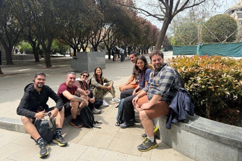 Eine Gruppe von Menschen sitzt auf einer Bank in einem Park mit Bäumen und einer Steinstruktur im Hintergrund. 2