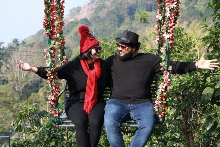 Two people sitting on a swing with red flowers, surrounded by greenery and hills. 3