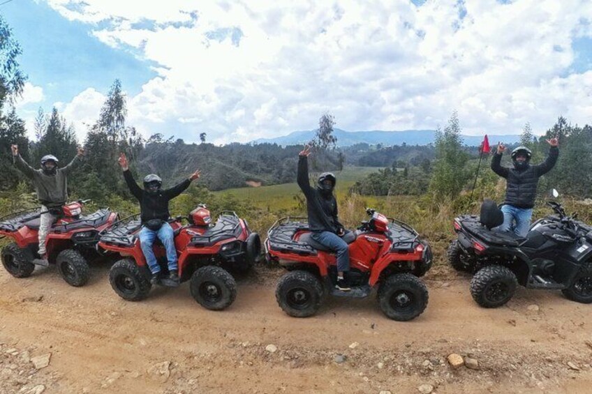 Vier Personen auf roten Quads mit erhobenen Armen, ein unbefestigter Weg und eine hügelige Landschaft mit Bäumen. 3