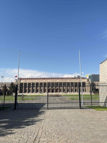 Asfaltoitu sisäpiha, jossa on aita, rakennus, jonka kyltissä lukee »Escuela Militar del General Bernardo O’Higgins», ja kirkkaan sininen taivas. 2