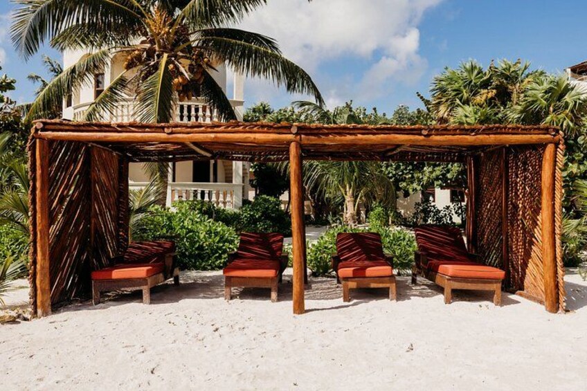 Een strand met een houten pergola, rode fauteuils met kussens, en een palmboom. 2