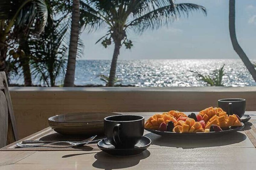 Un tavolo con un piatto di frutta, due tazze e una ciotola vicino a una finestra con vista sull'oceano. 4