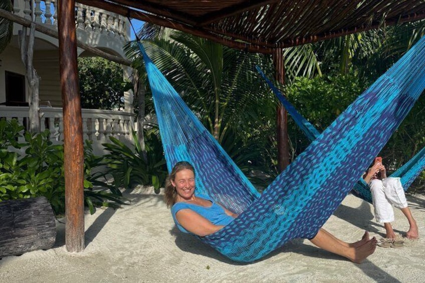 Una persona si rilassa in un'amaca su una spiaggia sabbiosa, con una casa e palme sullo sfondo. 2