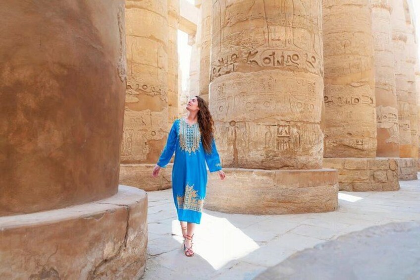 Una persona con vestido azul y bordados dorados caminando entre antiguas columnas de piedra con jeroglíficos. 2