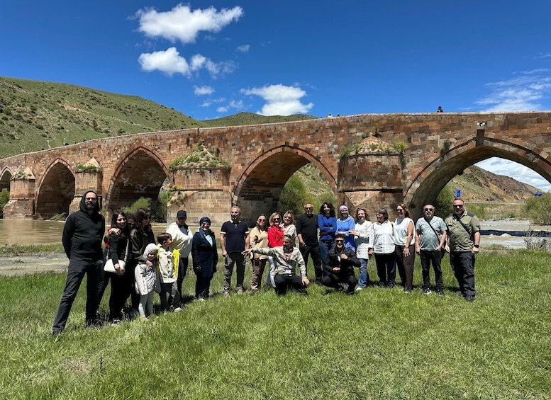 A group of people standing in front of an old stone bridge with arches over a river, surrounded by grass and hills. 4