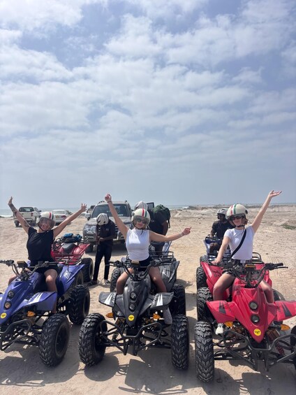 Three people on quad bikes on a sandy beach with vehicles in the background. 3