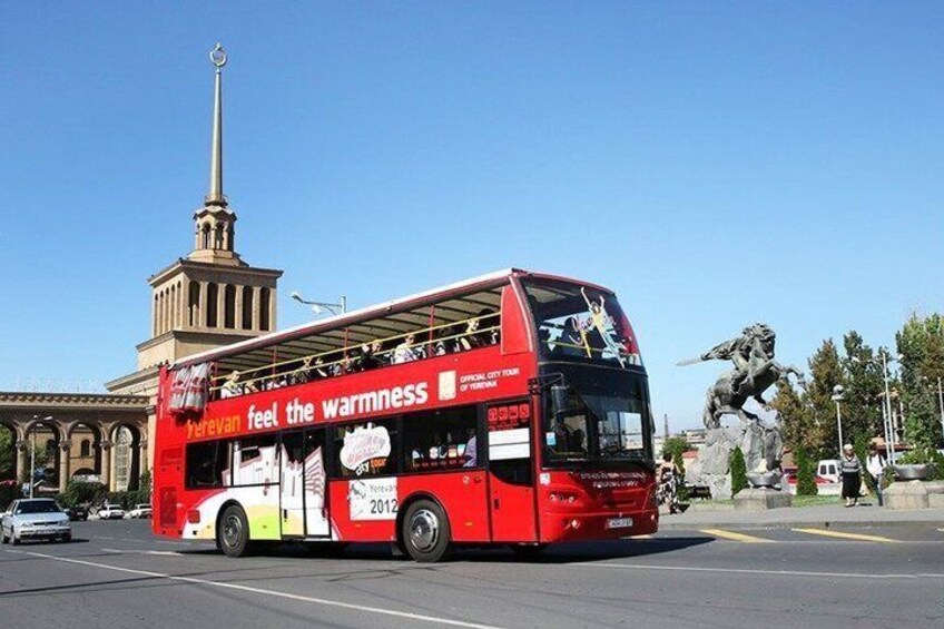 Ein roter, zweistöckiger Bus mit der Werbung 'feel the warmth', der auf einer Stadtstraße fährt, im Hintergrund ist ein Denkmal und eine Statue zu sehen. 1