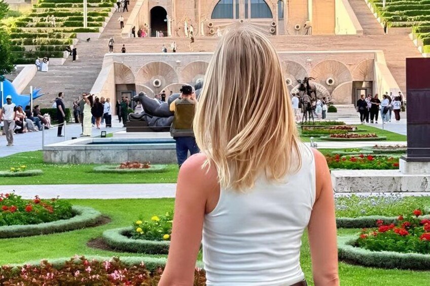 A woman in a white top facing a historic building with a fountain and landscaped gardens. 3