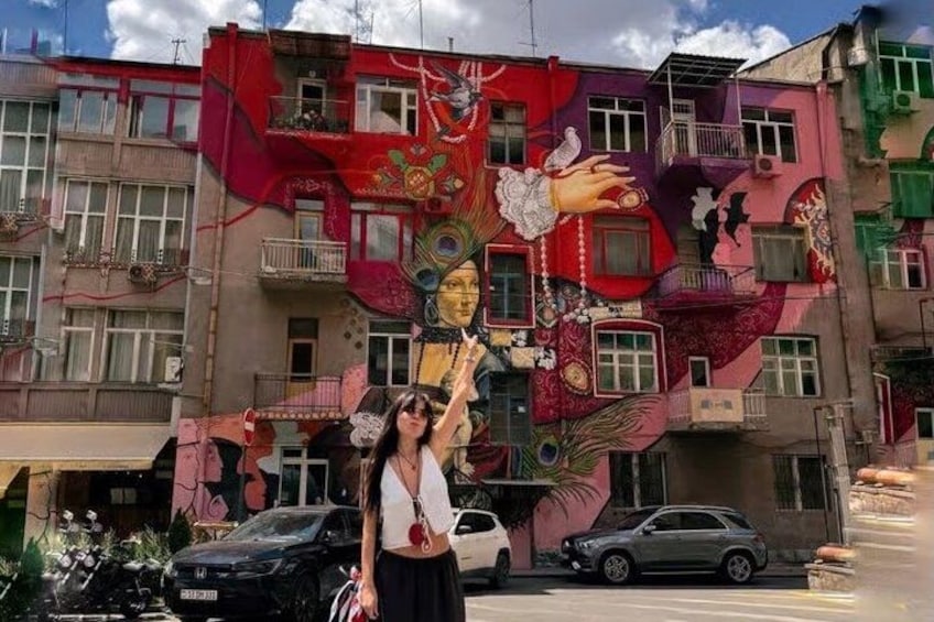 A colorful mural on a building, a person standing in front, and parked cars. 2