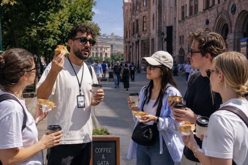 People standing on a street, some holding food and drinks, with a sign that reads 'COFFEE & GATA'. 1