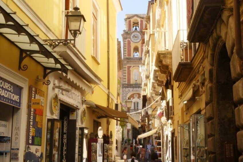 Una calle estrecha con comercios y una torre del reloj al fondo. 5