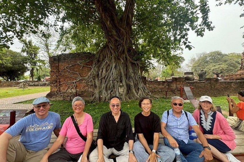 A group of people sitting on benches under a large tree with a brick wall and stone structures in the background. 2