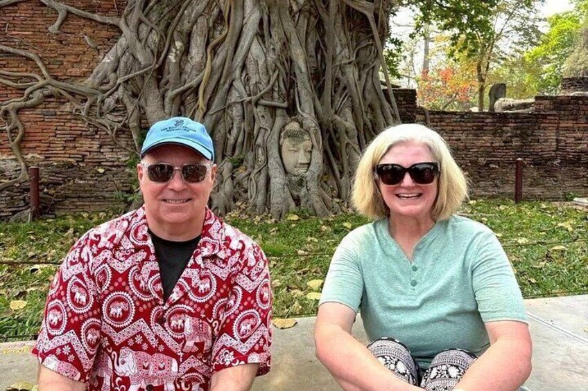 Two people sitting outdoors with a large tree and a brick wall in the background. 3