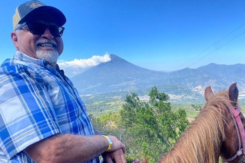 A man in a plaid shirt and cap, a horse with a pink halter, and a mountainous landscape with a volcano in the background. 2