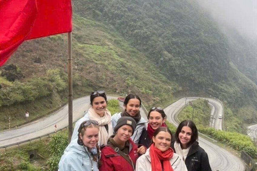 A group of people standing on a mountain road with a red flag, surrounded by lush greenery and a winding road. 1