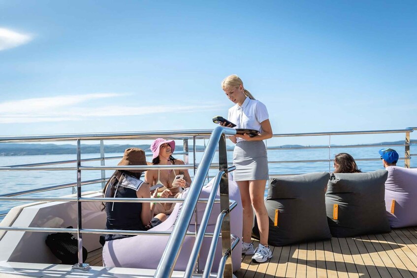 A group of people on a boat with a metal railing, a woman standing and holding a device, and others seated with cushions. 4