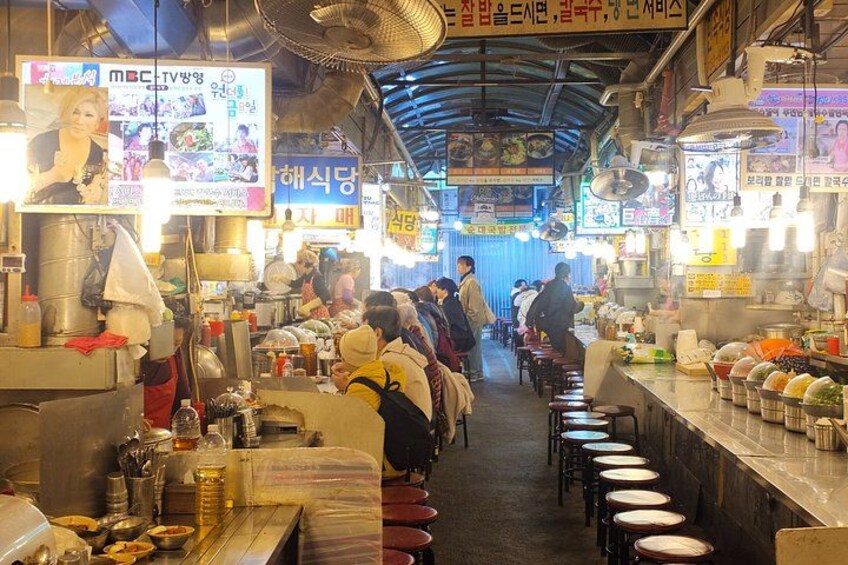 Un mercado cubierto animado con puestos de comida, gente comprando y diversos productos exhibidos. 2