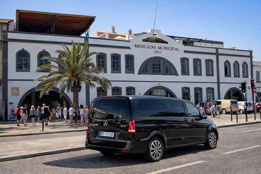 A black van driving past a white building labeled 'Mercado Municipal 1924' with palm trees and pedestrians in the background. 3