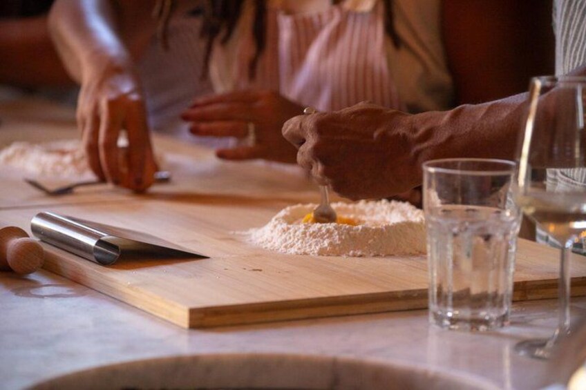 Une personne est en train de mélanger des ingrédients sur une planche en bois à l’aide d’un ustensile en métal. Un verre d’eau et un rouleau à pâtisserie se trouvent à proximité. 4
