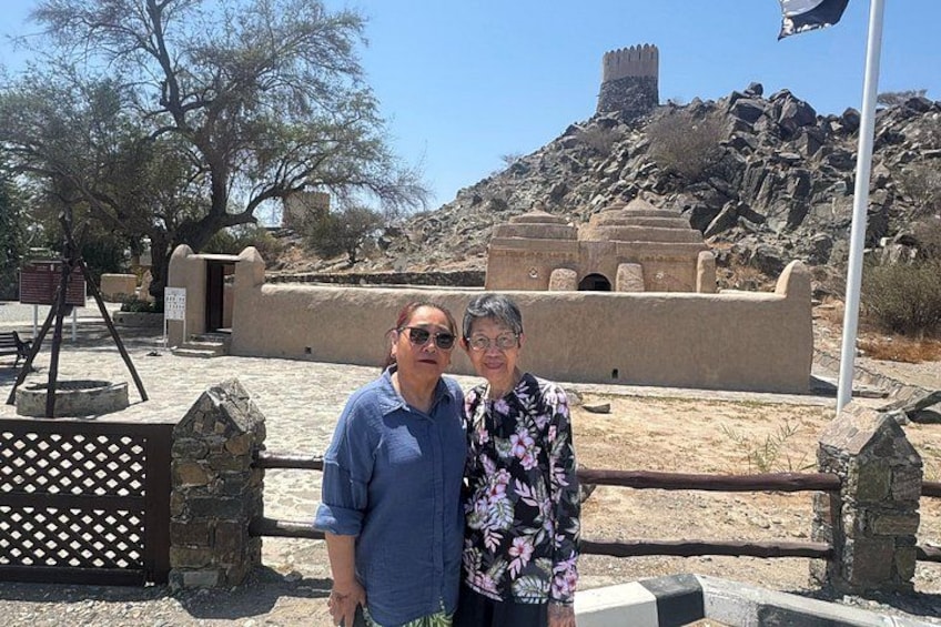 Two people standing in front of a historical site with a large stone structure and a flagpole. 1