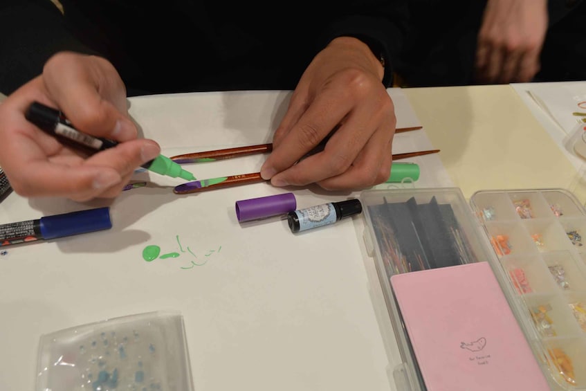 Hands holding markers and a pencil, with art supplies on a table. 3
