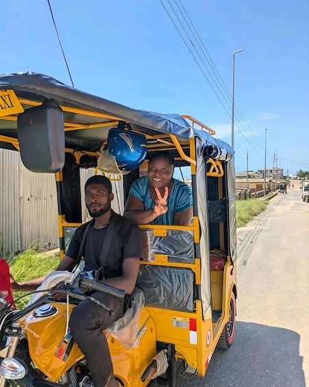 Two people in a yellow auto-rickshaw on a road with buildings and power lines in the background. 1