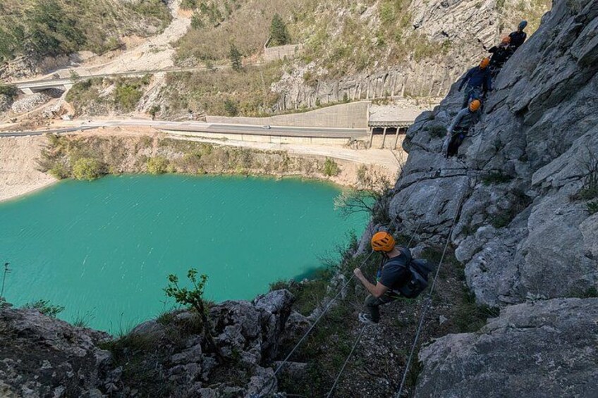 A group of people rappelling down a steep cliffside near a turquoise lake, with a bridge in the background. 4