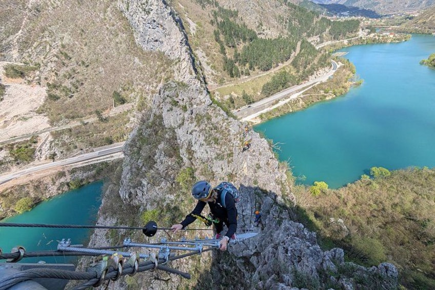 A person rappelling down a rocky cliff with a rope, overlooking a turquoise lake and a winding road. 1