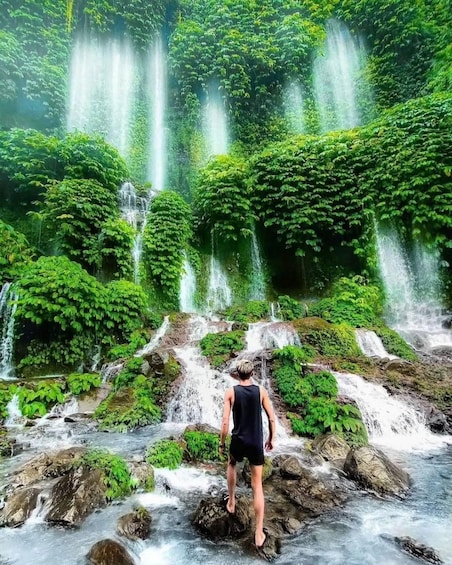 A person standing on rocks in a lush green forest with multiple waterfalls. 2
