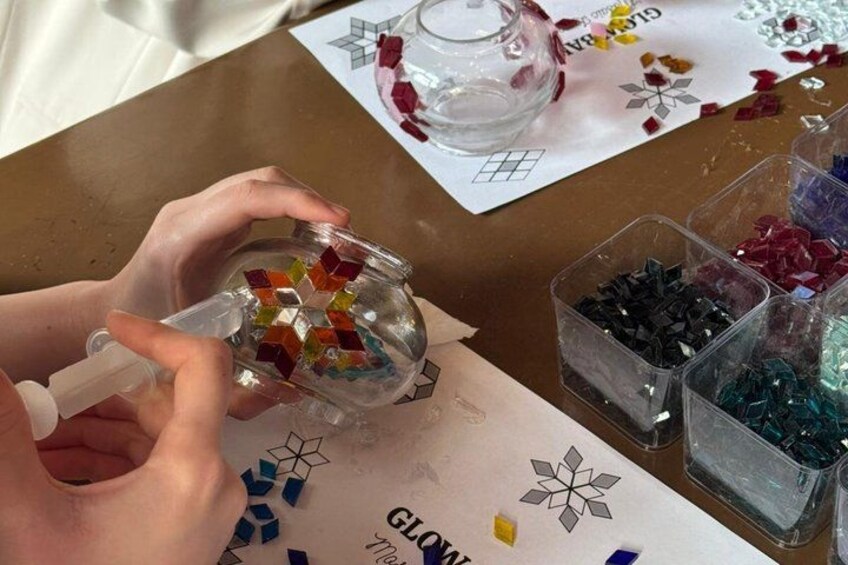 Händer håller i en glasburk med färgstarka mosaikbitar, en genomskinlig glasbehållare och små plastbehållare med olika färgade glasbitar, allt placerat på ett bord. 3